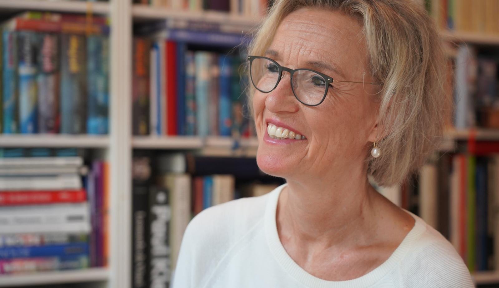 Claudia Peters lächelt und blickt zur Seite, hinter ihr ein volles Bücherregal.