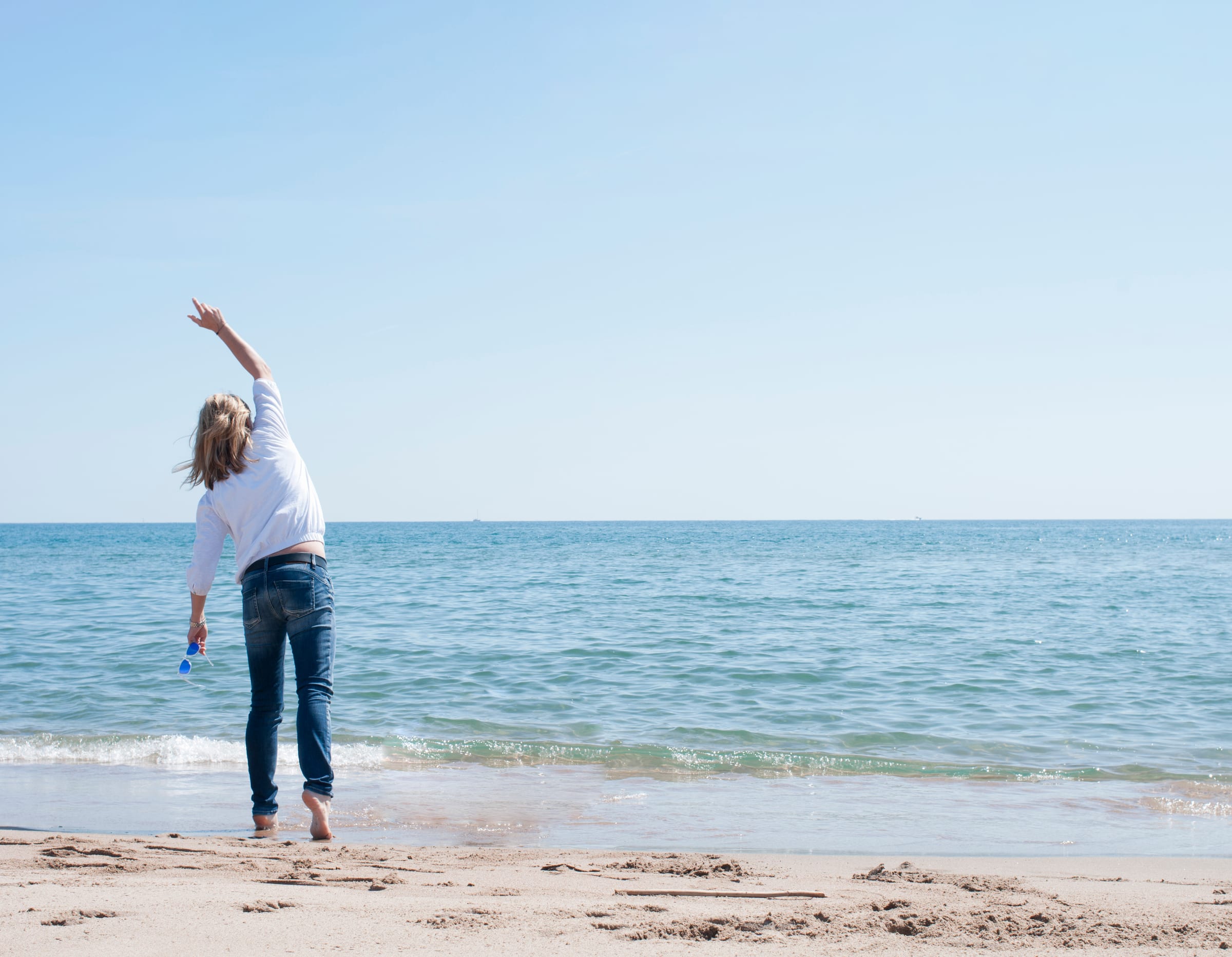 Eine Frau steht barfuß im flachen Wasser am Strand, von hinten gesehen, und streckt einen Arm nach oben.